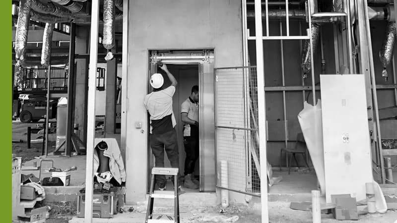 Construction workers installing commercial elevator door frame in unfinished building interior with exposed ductwork.
