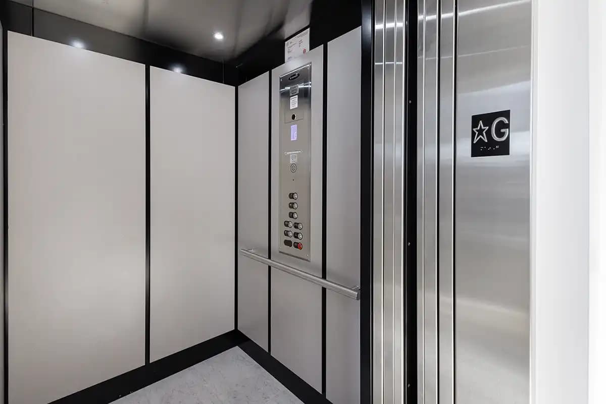 Savaria Orion LU/LA commercial elevator cab with white wall panels, black ceiling and trim, stainless steel handrail, car operating panel (COP), and automatic sliding doors