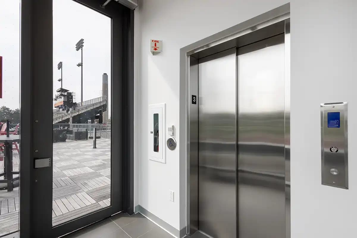 Savaria Orion LU/LA commercial elevator with stainless steel automatic sliding doors, digital hall call station, key switch, and fire safety equipment at sports facility entrance