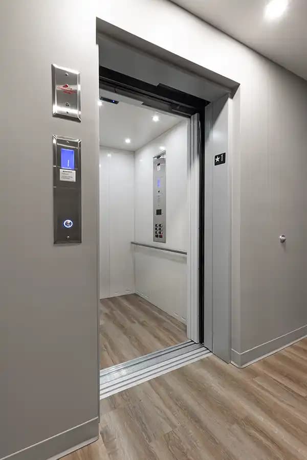 Savaria Orion LU/LA elevator cab with white steel wall panels, stainless steel handrail, automatic sliding doors, digital car operating panel (COP), LED ceiling lights, and wood-look flooring