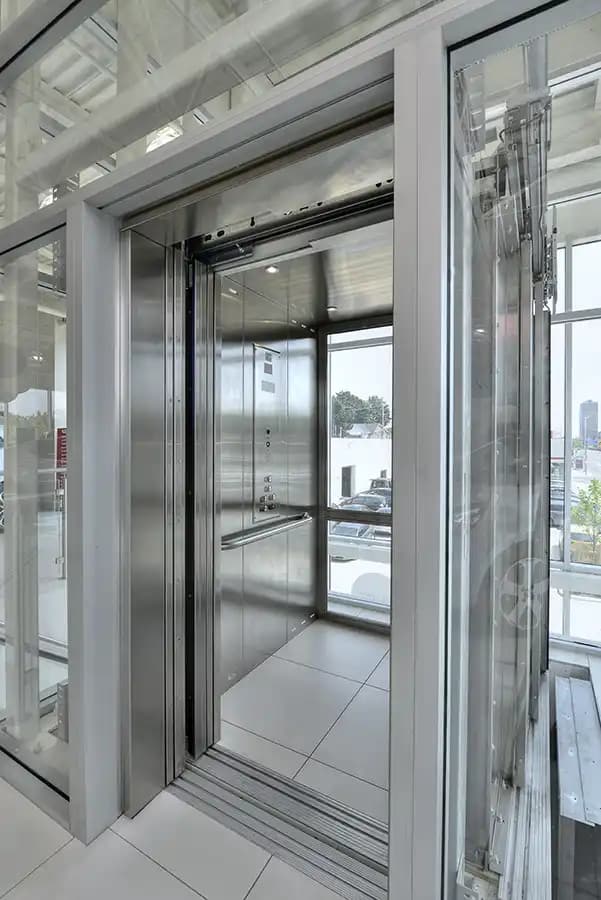 Savaria Orion LU/LA elevator with glass cab walls, stainless steel frame, automatic sliding doors, and handrail in modern retail building with natural lighting