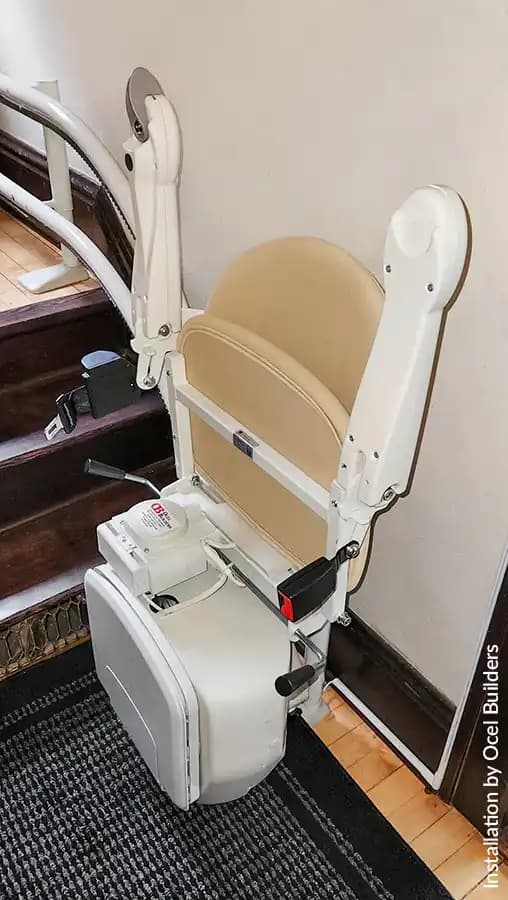 Savaria Handicare 4000 twin-rail curved stairlift with Simplicity Seat in sand upholstery, retractable seatbelt, charging station at bottom landing on wood stairs