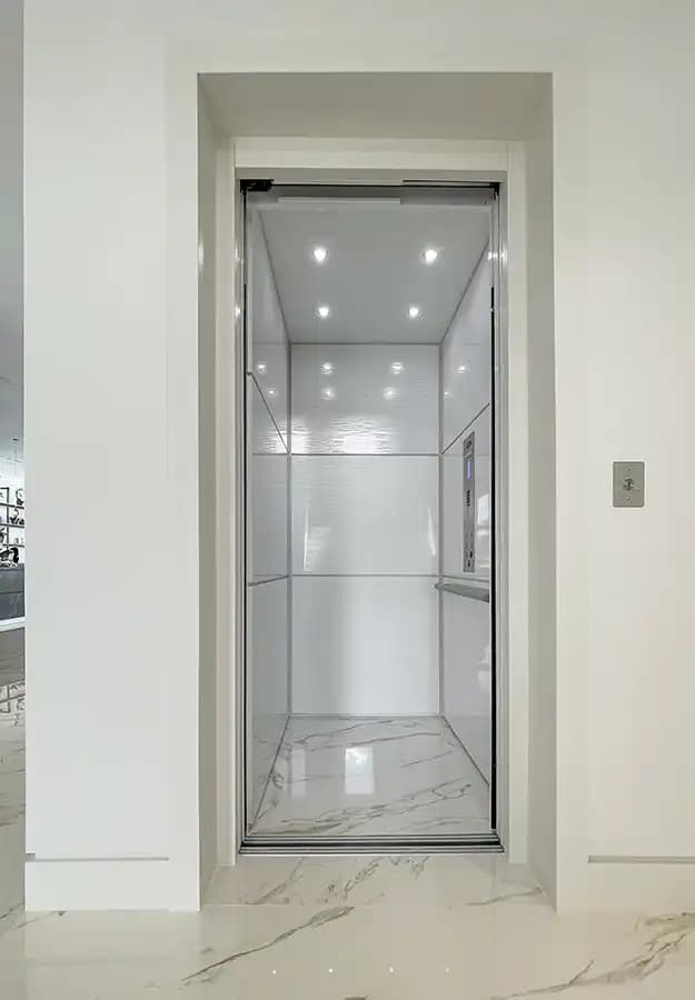 Savaria home elevator cab with white paneled walls, stainless steel handrail, recessed LED ceiling lights, and marble tile flooring